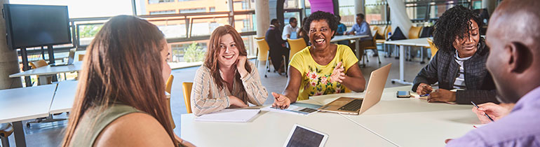 Postgraduate students conversing between themselves over a white table, using tablets and laptops for study