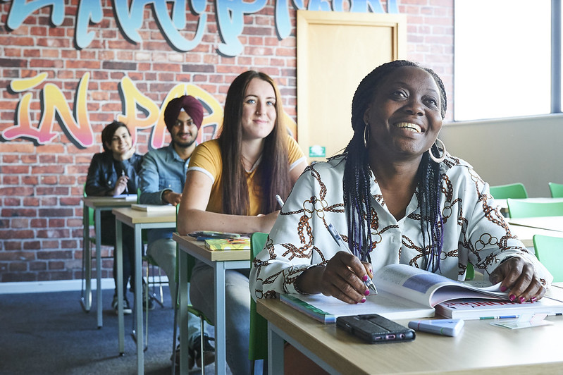 Students at desks in a classroom, holding textbooks open and facing towards the front of the room