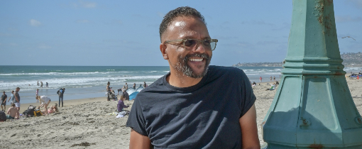 David Odusanya wearing a black t-shirt, standing against a lamppost in front of a beach