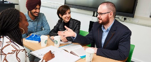 Photograph of a teacher and students addressing each other around a table, with coffee cups and books arranged between them.