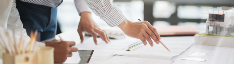 Image of various plans on paper arranged on a table-top, being surveyed by a person holding a pencil to the paper.