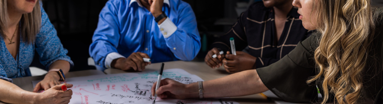 Photograph of four people conversing across a table with a whiteboard between them
