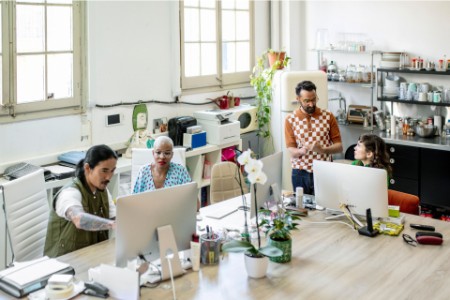 Photograph of four people working in an office, a large table hosting two Mac computers, kitchen appliances arranged on shelves behind the workspace.