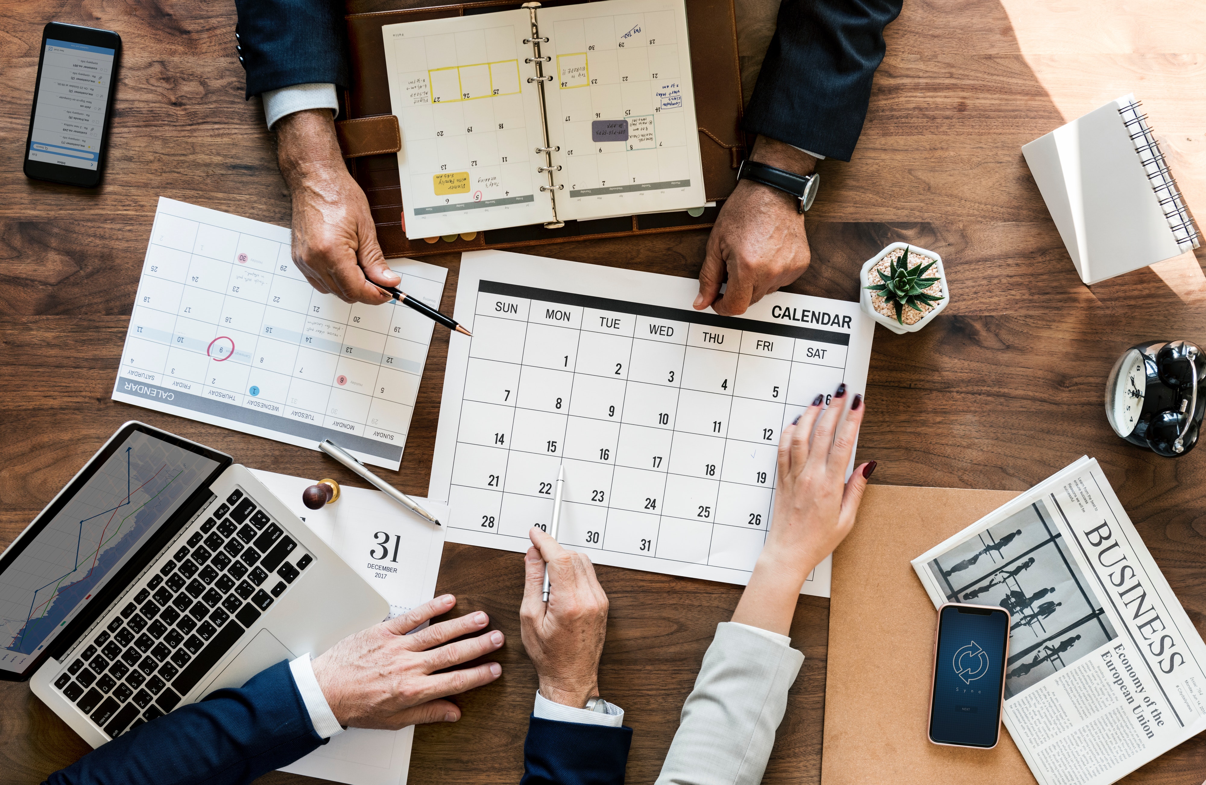 Image of several pairs of hands working on a table-top with notebooks, calendars and laptops.
