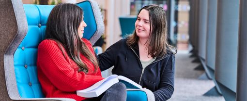 Two female students looking at each other one sitting down with book and the other crouched down