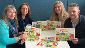 Four people at a table holding up posters for the Finding our Funny Roots project
