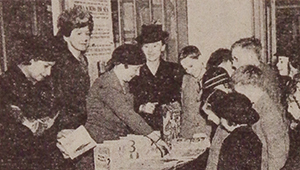 Black and white photograph of a crowd of people, including Beatrice Warde, surrounding books on a table