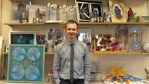 Steve Sharp wearing a button-down shirt and tie, standing in front of and to the side of framed artwork on tables and shelves