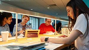 Four students sitting at a lit table, working with pen and paper or digital tablets