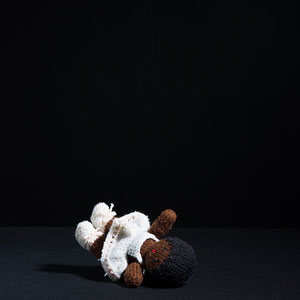Photograph of a knitted doll wearing a ballet outfit, left on its back in a black room.