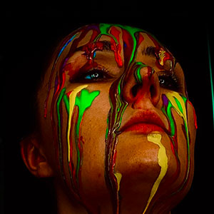 Picture of a person with their head raised upwards, various coloured paints streaming down their face.