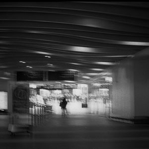 Blurry black and white photograph taken inside a train station, billboards visible above a doorway with a figure travelling through it.