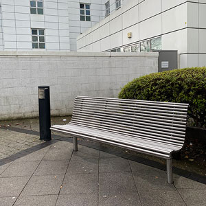 A silver metal bench on a footpath outside a large grey building.