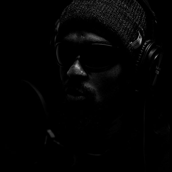 Black and white portrait photograph of a man wearing dark glasses, a beanie and headphones.