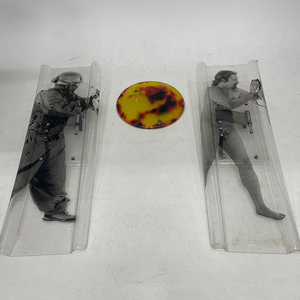 Images of two people facing right printed on separate plastic slides, one wearing a protective helmet and mask, the other undressed, between them a circle with a printed fiery texture.