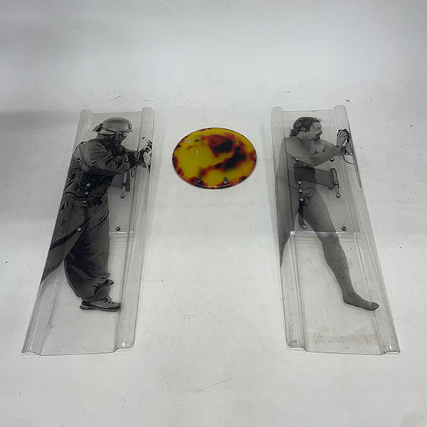 Images of two people facing right printed on separate plastic slides, one wearing a protective helmet and mask, the other undressed, between them a circle with a printed fiery texture, photographed from afar.