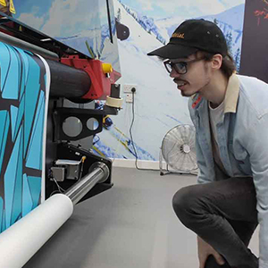 Illustration student, Patrick Campbell-Brookes, photographed in the School of Creative Industries Print Support Hub. Patrick is knelling on the floor watching his large scale artwork printed on one of the large format colour printers in the room. He is wearing glasses, a baseball cap and a denim jacket.