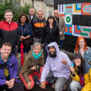 Fine Art Staff and students posing for a group photo in front of a brightly painted sign, with multicoloured text on a black background, saying: 