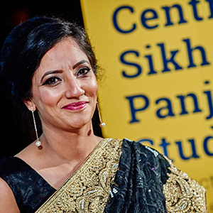 Dr Opinderjit Kaur Takhar MBE. Dr Opinderjit is wearing a traditional black Sikh dress with a gold sash. She is photographed against a yellow banner that says: Centre for Sikh and Panjabi Studies.