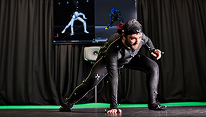 Male student using the motion capture facilities in our Screen School. They are wearing a special suit with sensors that records their movements. They have their legs apart with one hand pressed against the floor whilst the other is resting on their knee