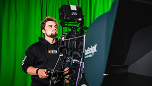 Male Film and TV Production student using a large video camera in the TV studio. You can see a green screen in the background