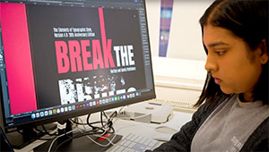 Female Graphic Design student designing a magazine front cover, using one of the Apple Macs in our Graphic Design studios. The screen is showing a close up of the cover which has large text that says:
