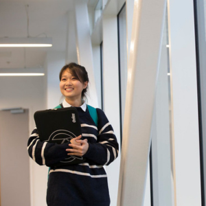 A student with a laptop bag between their arms standing next to a window