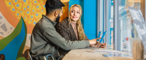 Two students with laptops at a desk near a window, looking at each other and conversing