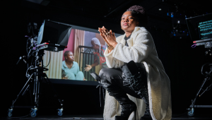 A person photographed from a low angle in a dark room, surrounded by camera equipment.