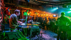 A club with a band performing on stage, a drummer and two guitarists playing under florescent green lights