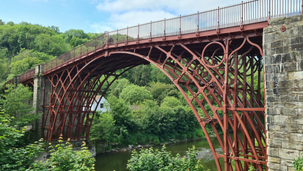 A fenced iron bridge crossing a body of water, comprising an ornate arch stylised with chain shapes, a house visible between the trees on the opposite side of the bridge
