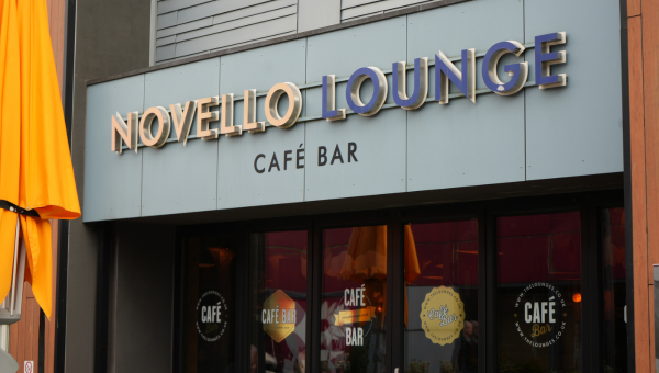 Exterior of Novello Lounge Café Bar, with prominent signage on and above the tall glass windows, a parasol for an outdoor seating spot visible