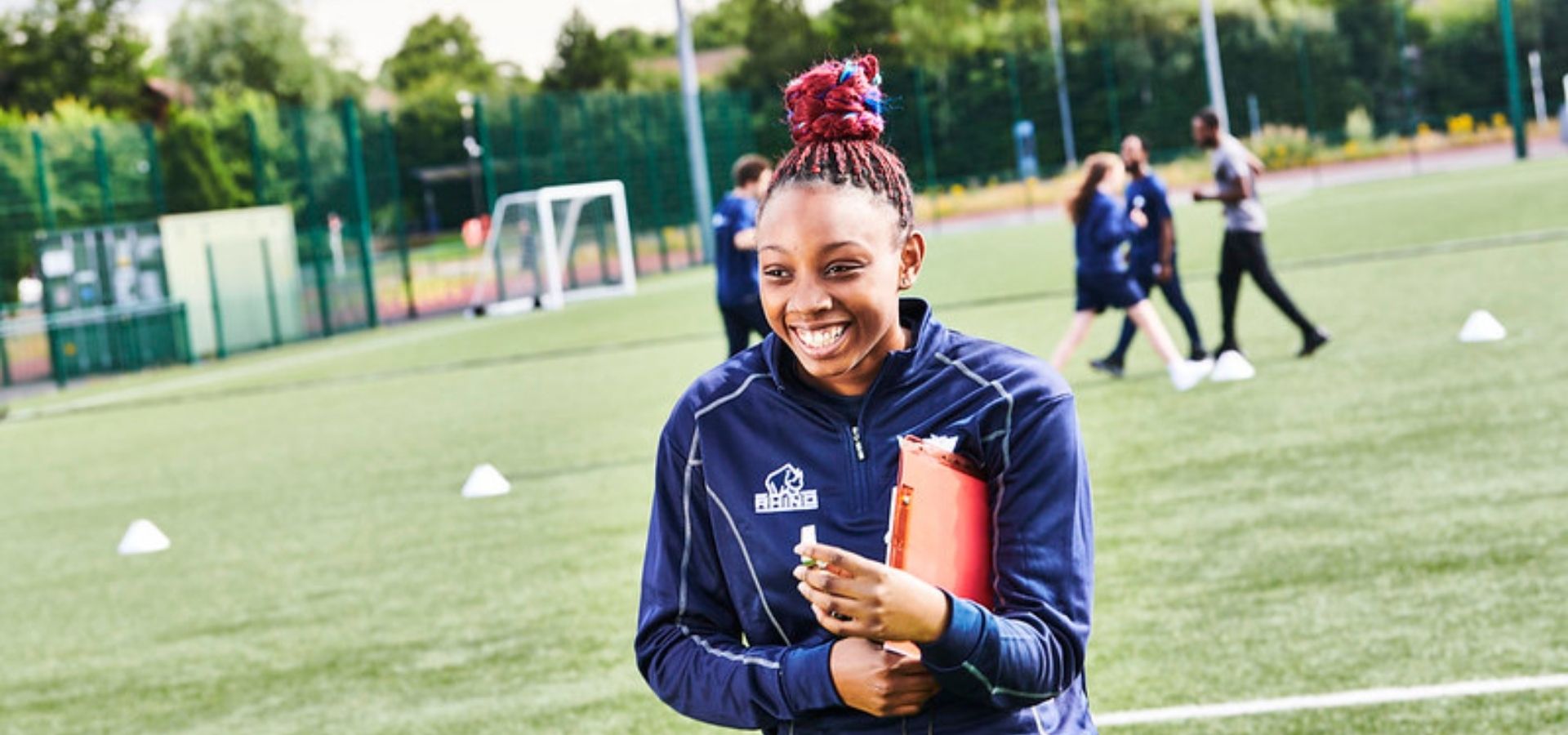 Student on football pitch at Walsall Campus, in uniform and holding a clipboard, with four students in the background behind her