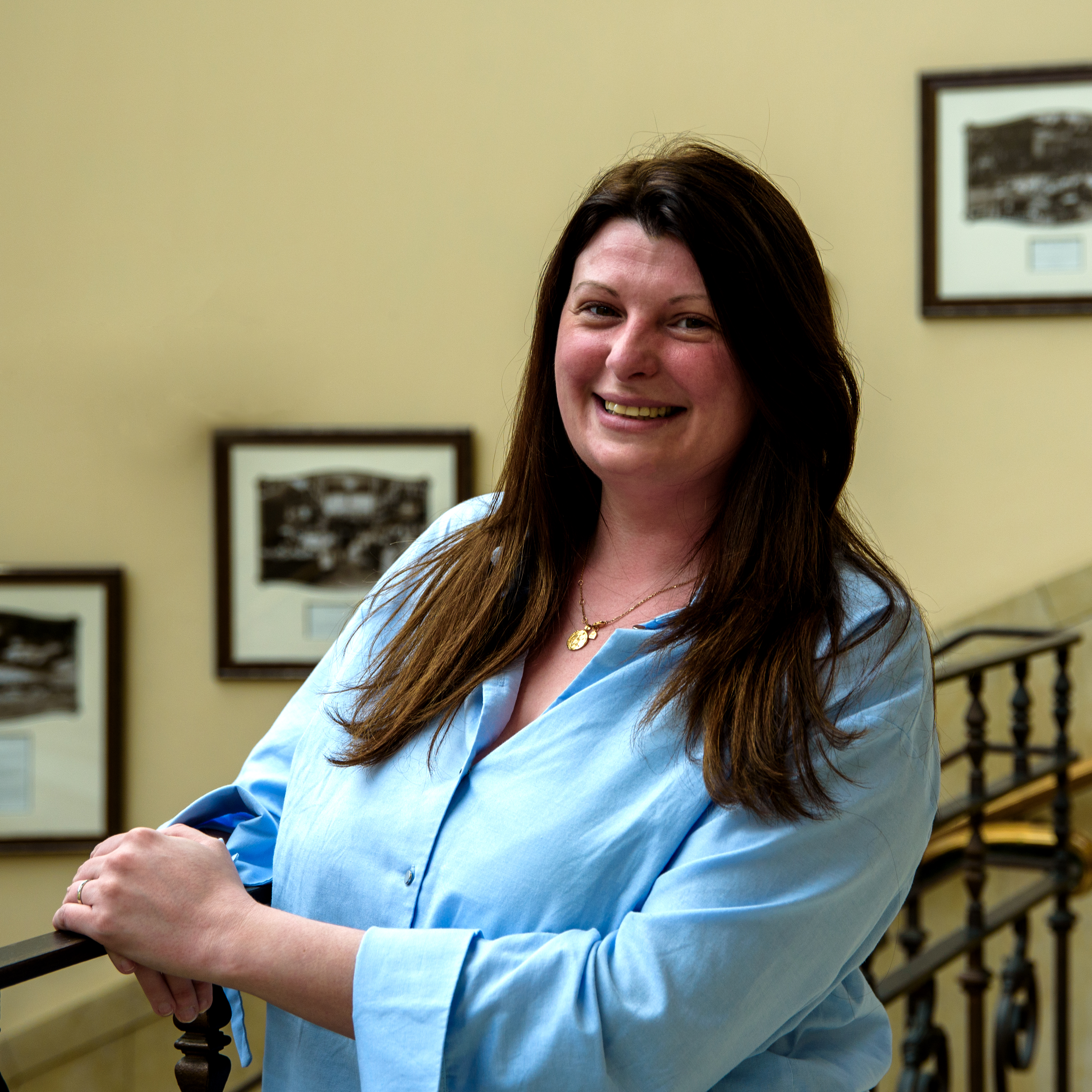 A white woman in her 30s with long brown hair, standing in front of a staircase