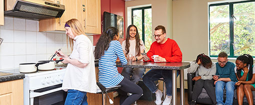 A group of students in a communal kitchen area, sitting around a table or on a sofa, one student working at the hob in the kitchen