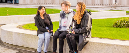 Three students, one male and two female sitting chatting in the courtyard at the University of Wolverhampton's City Campus in the sunshine.